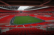 The interior o a empy stadium as viewed frae its upper tier o seatin. The seats is a vivid red an the pick is a vivid green. The pale grey sky is visible throuch a openin in the ceilin abuin the pick.