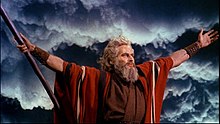 A color screenshot of a man with a beard wearing a robe. He has his hands raised at his side, and his right hand is holding a staff. Dark clouds can be seen in the background.