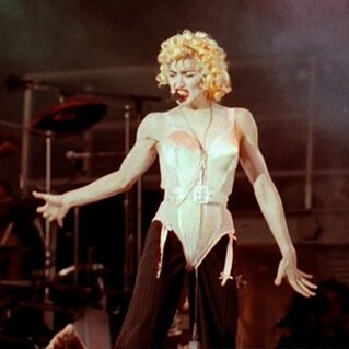 The image of a young blond woman. She is wearing a salmon corset. Her hair is short and curly. She has bright red lips and appears to be singing to the audience on her left while looking down.