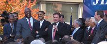 A crowded stage of politicians both white and African American stand before supporters and press.