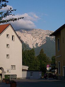 Une montagne, se faisant recouvrir d'une nappe de nuages à l'arrière plan, est limitée par des habitations et de la végétation au premier plan.