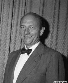 Photo en noir et blanc. Un homme debout, portant un veston, sourit.