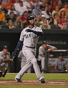 An dark skinned man wearing a gray baseball uniform with "SEATTLE" on the chest stands holding a baseball bat in his right hand as if having just taken a left-handed swing.