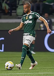 Um homem jogando futebol, se preparando pra dar um passe com o pé direito, com feição de concentrado.