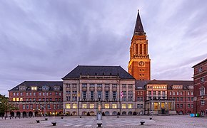 Grand bâtiment rectangulaire avec un petit toit, flanqué sur le côté d'une tour pointue.