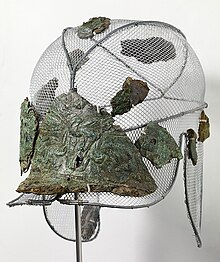 Photographie d'un casque fragmentaire orné de motifs végétaux, en fer et bronze, de couleur verte.
