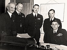 Cinq hommes, dont quatre debout et un assis, portant des uniformes militaires de couleur foncée.
