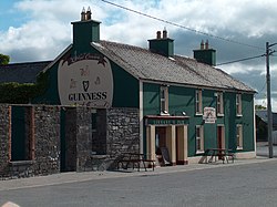 Photo of Linnane's Pub - A green colour building with "Linnane's Pub" written in light yellow above the main door. The door and window frames are light yellow. There is a large advertisement for Guinness on the side of the building. There are two park benches in front of the pub. There is a grey structure on the left side of the pub. The photo was taken during the day when the sky was blue with white clouds. It is taken from outside, and from opposite side of the road