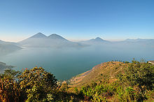 Vista panoràmica entre els turons sobre un gran llac banyat en una lleugera boira. Les ribes del llac es corben des del primer plànol a l'esquerra cap enrere i cap a la dreta, amb tot de volcans a la riba oposada, emmarcat per un cel blau clar.