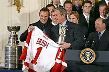 Photographie de Yzerman à côté de la coupe Stanley et qui remet à Georges W. Bush un maillot des Red Wings avec le numéro 1 et son nom.