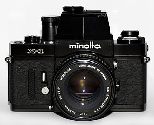 A black-finished single lens reflex camera made by the Minolta Corporation; this camera is equipped with a normal lens of 50 mm focal length and a distinctive viewfinder with a vertical line bisecting its profile which provides aperture-priority autoexposure.
