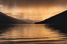 Dans une atmosphère ocre très contrastée, un ciel couvert et tombant, un lac d'eau calme, bordé de noir et de deux collines.