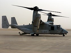 A side view of an MV-22 resting on sandy ground with its ramp lowered.