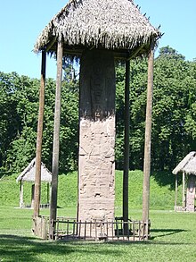 Un monumento alto y estrecho cubierto por un techo de paja. Dos estelas más visibles en el fondo, también están cubiertos por techos de paja, en un fondo de árboles.