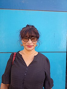 Oberkörper einer Frau mit schwarzem Oberteil und Sonnenbrille, die frontal zum Fotografen steht und breit lächelt. Im Hintergrund eine blaue Wand.