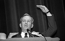 Photographie en noir et blanc d'un homme en costume et faisant un discours devant des micros