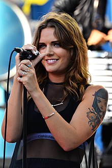 A woman with brown hair wearing a black top and holding a microphone.