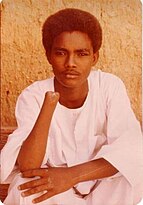 shown is Abdul Rahman Ali, a young Sudanese dressed in a jalabiya, a traditional Sudanese white garment, sitting against a textured, warm-toned wall. Ali has a calm and contemplative expression, resting their left arm on their lap while their hand supports their face. Ali's right arm is amputated just below the elbow.