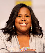 Porträtfoto einer sitzenden Amber Riley. Sie trägt große, goldfarbene Ohrringe, ein fliederfarbenes Kostüm und eine leicht gewellte Bobfrisur.