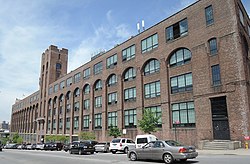 View from Lafayette Avenue showing the rows of windows surmounted by brick arches and set back from the face of the building. A brick tower is in the middle of the façade. The street slopes away from the viewer, exposing additional rows of windows.