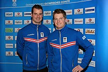 Photographie de deux hommes portant la même veste de survêtement bleue à parements rouges et blancs, posant devant un panneau publicitaire.