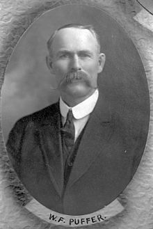 A torso shot of a middle-aged white man with receding dark hair and a large moustache, wearing a three piece suit