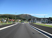 Foto einer Straße, die in einem Ort auf einen Kreisverkehr hinführt, dahinter ein Berg