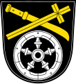 Gemeinde Illesheim In Schwarz über einem fünfspeichigen silbernen Rad schräg gekreuzt ein gestürztes goldenes Messer und ein goldener Kreuzstab.