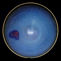 A ceramic bowl is viewed from directly above. The multitoned blue glaze has a crazed and mottled finish, darker near the rim of the bowl, and lighter in the centre. The rim itself is yellow. An off-centre patch of purple and red interrupts the blue finish.