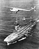 An aircraft carrier underway, with two helicopters flying overhead