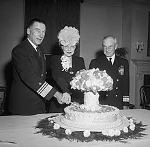 Un hombre con uniforme naval y una mujer con sombrero cortados en un pastel con la etiqueta Operación Cruce de caminos, y con la forma de una nube en forma de hongo, mientras otro oficial naval observa.