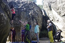 Mehrere Personen zwischen Sandsteinblöcken; eine Kletterin an einer Wand, ca. 2 m über dem Boden; ein Mann hat beide Arme in Richtung der Kletterin ausgebreitet