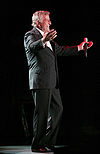 An older man holding a microphone in one hand, his arms held out, smiling and wearing a black suit with a white dress shirt.