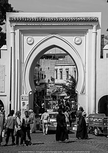 Photographie en noir et blanc d'une grande porte extérieure blanche aux formes orientales. Plusieurs personnes et véhicules entrent et sortent par la porte.