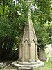 A stone drum on a stepped base below a tapering section, then a decorated box, then a cross. Around the drum are arched panels with carved writing. It stands in front of trees, some of whose branches partly obscure the decorated box. To the right is a gravestone.