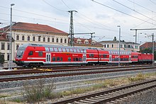 Ein rot lackierter Zug aus einem Doppelstocksteuerwagen, einem Doppelstockmittelwagen und einer Lokomotive. Auf dem Mittelwagen befindet sich ein weißer Schriftzug „VVO“. Im Hintergrund ein Hotelgebäude.