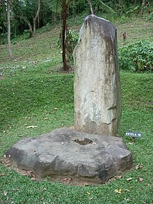 A tall, narrow standing stone set in a grassy area with an overgrown mound in the background. A flat circular stone lays at the base of the standing stone, it has been carved into a rough cogwheel shape.