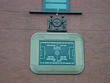 Một bia đá khắc hình ảnh của một sân bóng đá và một số tên cầu thủ. Nó được bao quanh bởi một đường viền đá trong hình dạng của một sân vận động bóng đá. Phía trên bảng là một tấm gỗ khắc hai người đàn ông cầm một vòng hoa lớn.