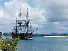 A three-mastit sailin ship wi its sails furled, sittin motionless on a body o watter. A beach an forestit hills are in the backgrund.