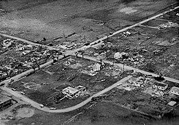 Aerial view showing wreckage, debris, and dismantled homes with farmland in the background