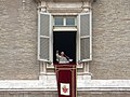 Farbfotografie von Papst Benedikt 16, der an einem durchsichtigen Rednerpult am offenen Fenster steht und seine rechte Hand hebt. Am Fenster sind graue Fensterläden und ein dunkelroter langer Stoff mit goldenen Verzierungen und das Wappen hängt am Fenstersims.