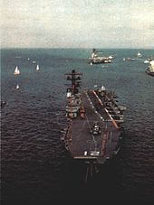 Colour photo of a small aircraft carrier, taken from an elevated position. The carrier is sitting bow on, with one of her anchor chains deployed.