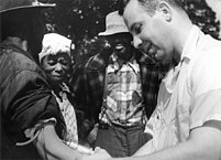 A white doctor has inserted a large needle to draw blood from a black man who stands with his arm at his side. The photo is in black and white. The black man is on the right while the white man stands to the left of him in the frame. Two other black Americans gaze down at the black man's arm as his blood is drawn.