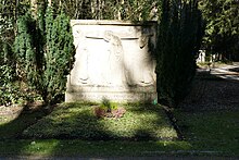 Von der Sonne beleuchteter, rechteckiger Grabstein mit einem Relief von drei Menschen, die einen vierten zu Grabe tragen. Darunter die Namen Mary von Stuck und Franz von Stuck. Links und rechts vom Grabstein dunkel Büsche, davor das mit Bodendeckern und Erika bewachsene Grab.