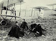 Photographie noir et blanc de quatre hommes assis dans l'herbe, en uniforme, devant un biplan.