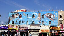 View of mural seen over small store front businesses. Mural has a woman whose thoughts appear as text in a cloud saying "History is myth."