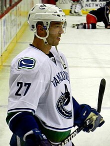 An ice hockey player seen from the side in a ready position. His legs are planted as he holds his stick outward in front of him. He wears a white jersey with blue and green trim.