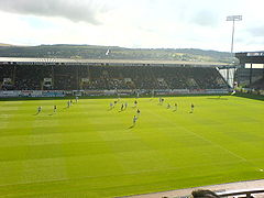 A football stand photographed from another stand, while some footballers are on the pitch