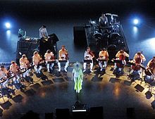 Image d'un concert présentant une femme masquée entouré par un ensemble d'instruments à cordes, un homme sur un ordinateur et un homme sur batterie