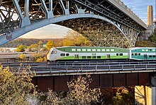 A green-and-white GO train passing under a large arch bridge in Hamilton, Ontario.